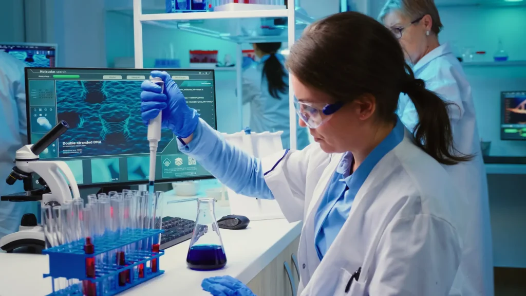 A scientist is using a pipette in a lab with test tubes and a computer showing DNA.
