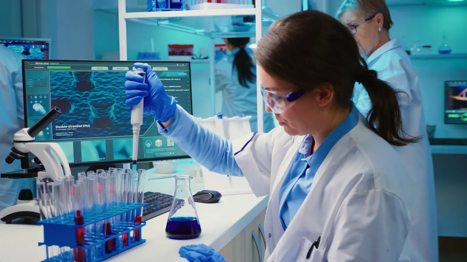 A scientist is using a pipette in a lab with test tubes and a computer showing DNA.