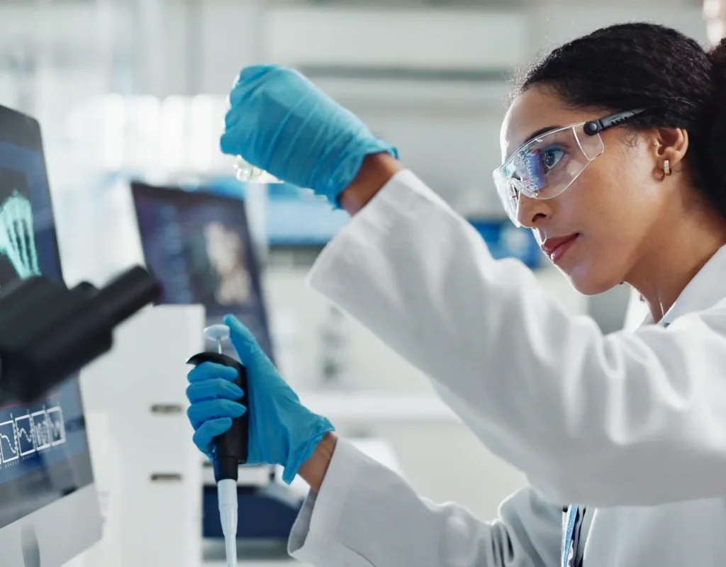 A female scientist in a lab coat, gloves, and safety glasses works with a vial and pipette in a lab.