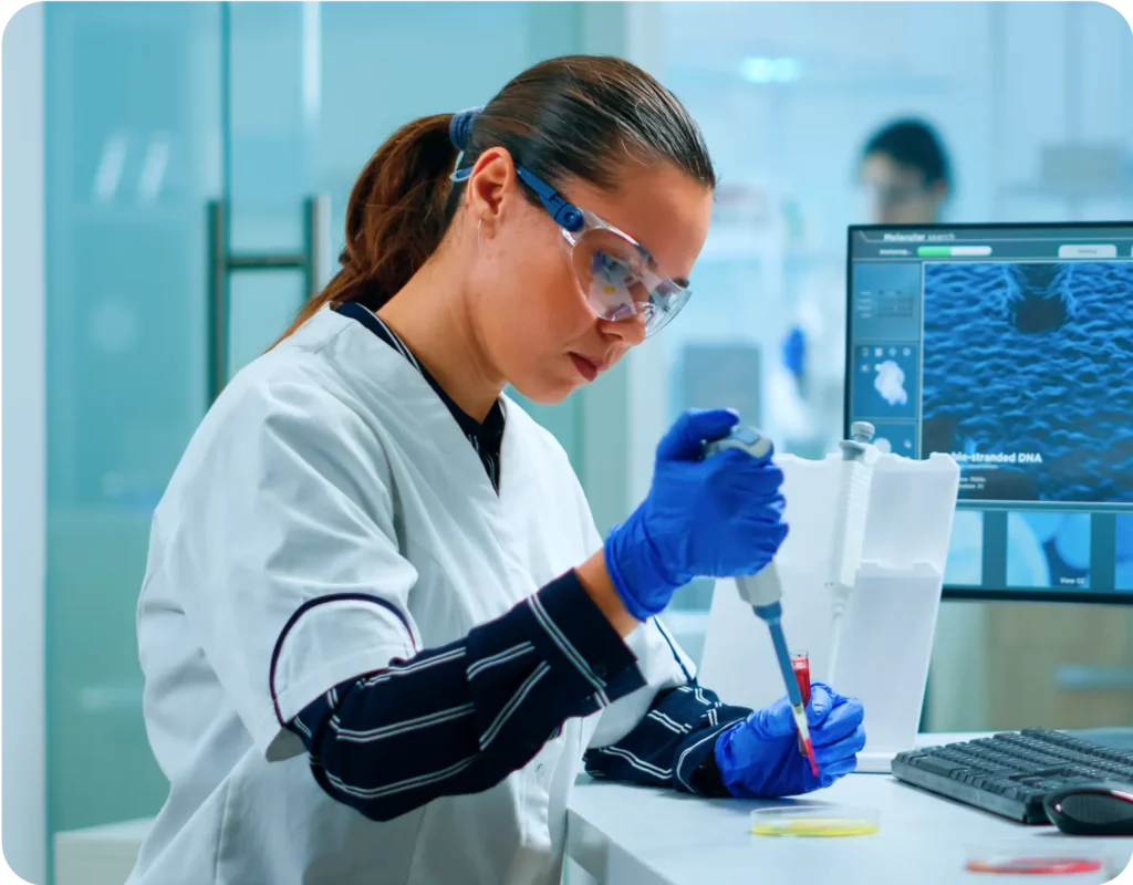 A female scientist in a lab coat and gloves uses a pipette in a lab, with a monitor showing DNA data